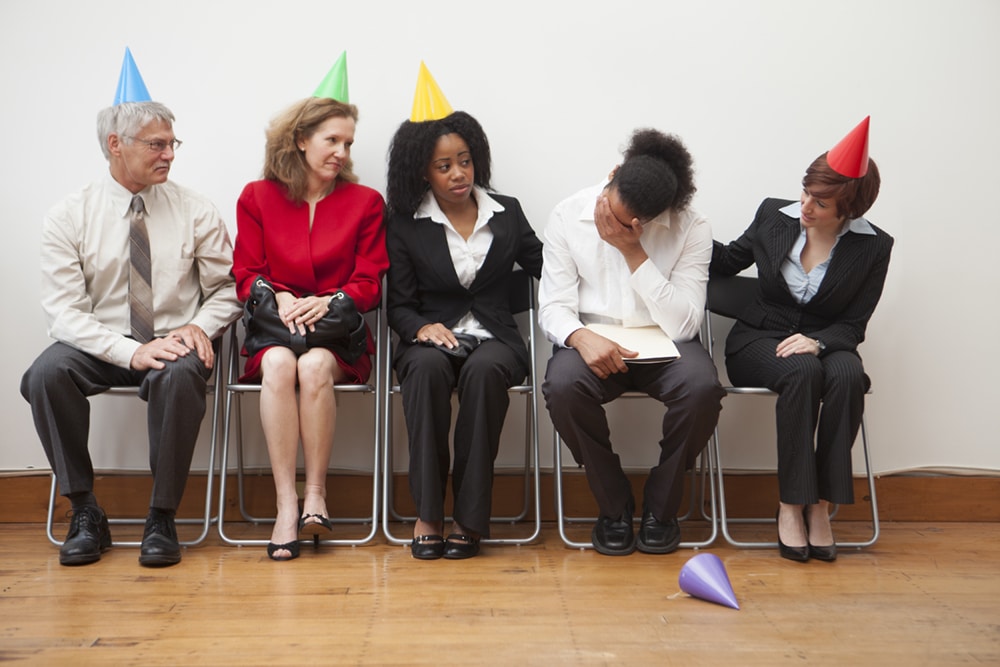 A group of office workers sitting awkwardly by the wall in a party, wearing multi-colored party hats.